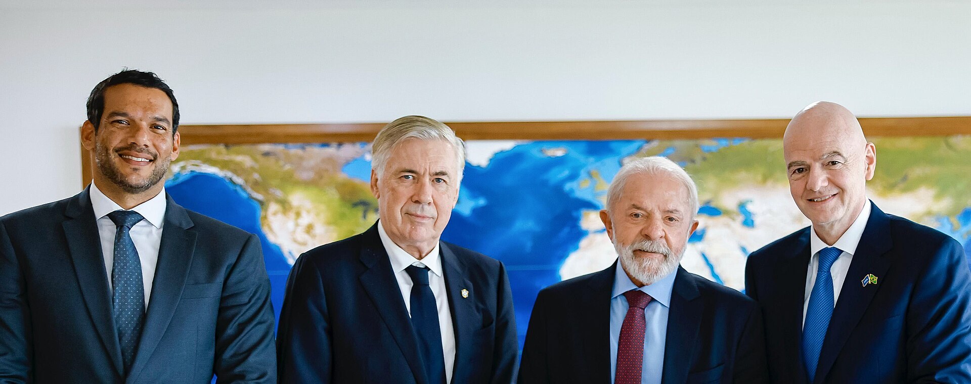 Four men in suits pose in front of a world map; one with gray hair holds a soccer ball