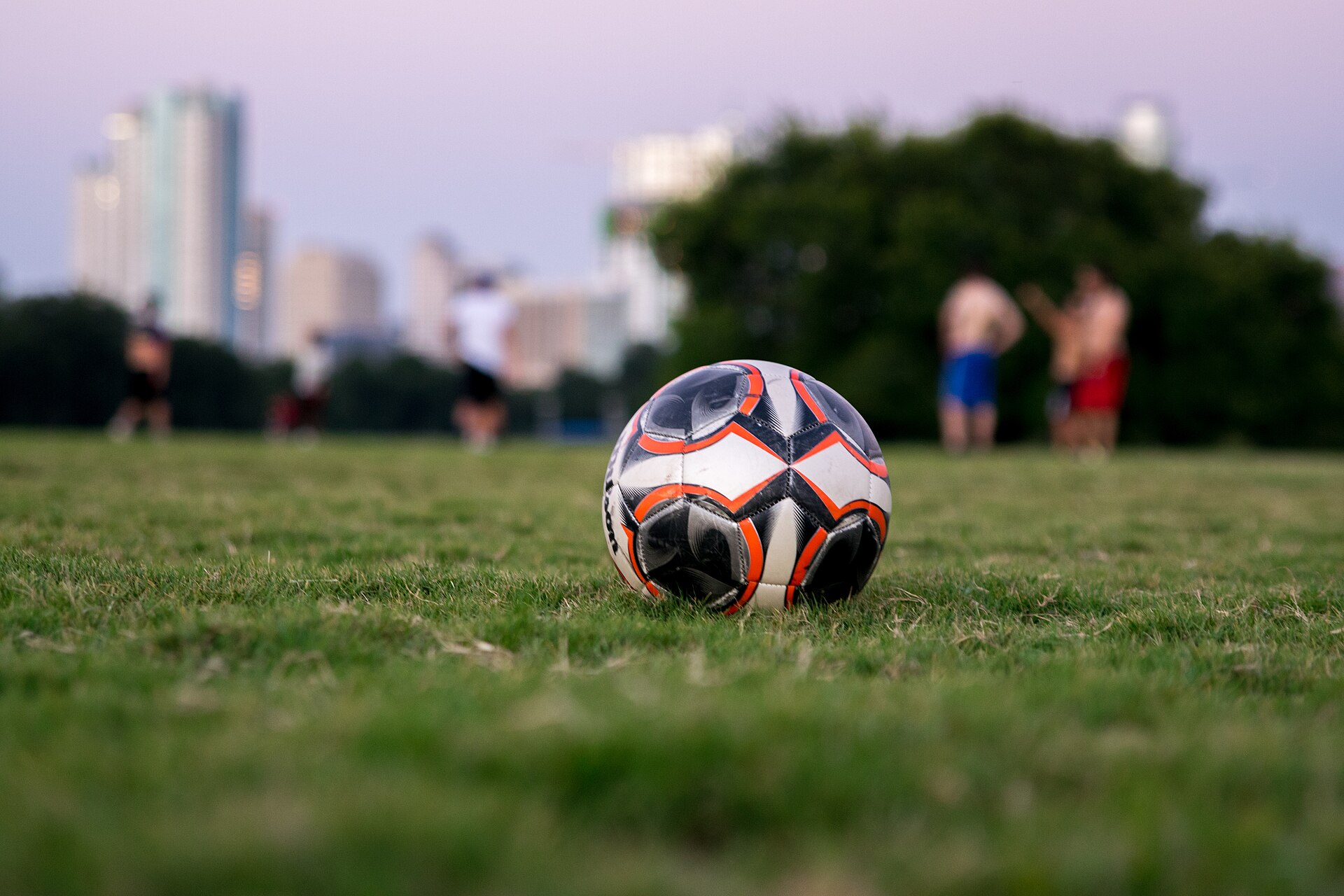 A white, black and orange soccer ball on the grass, with blurred players in the background