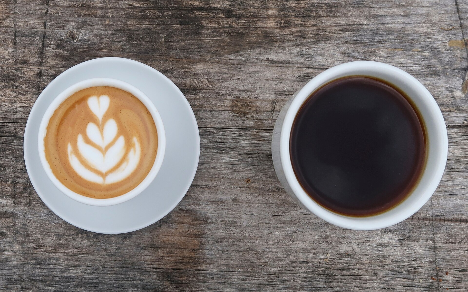 A cup of latte with a leaf design in the foam and a cup of black coffee, seen from above on a wooden table