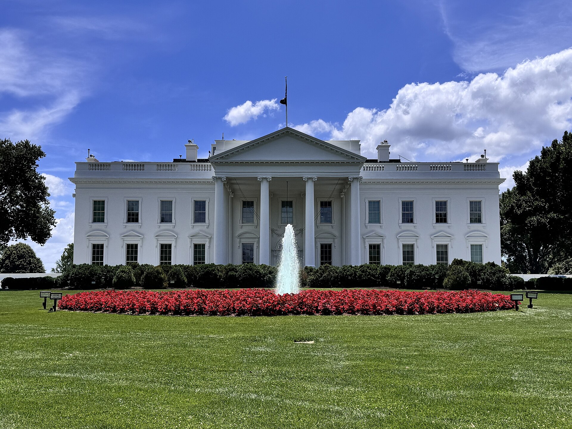 The white columned facade of the White House, with a fountain and a bed of red flowers in front, under a blue sky