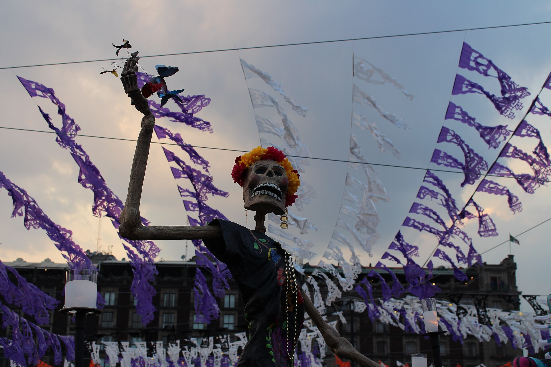 Uma catrina gigante com coroa de flores levanta um braço entre tiras de papel picado roxo e branco