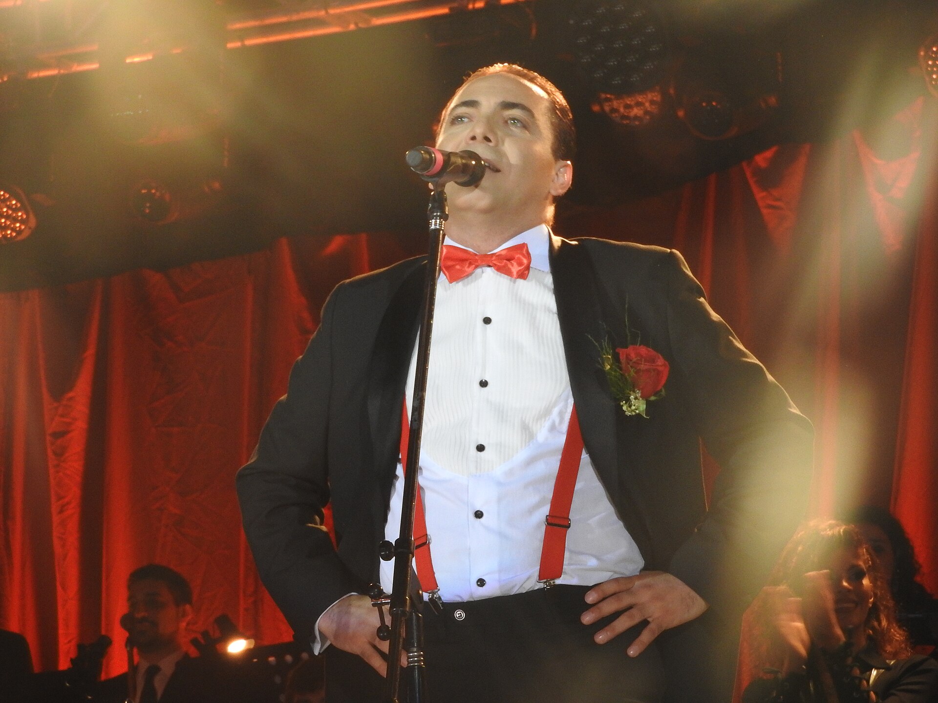 Cristian Castro canta diante do microfone de smoking preto, gravata-borboleta vermelha, suspensórios vermelhos e uma rosa vermelha na lapela