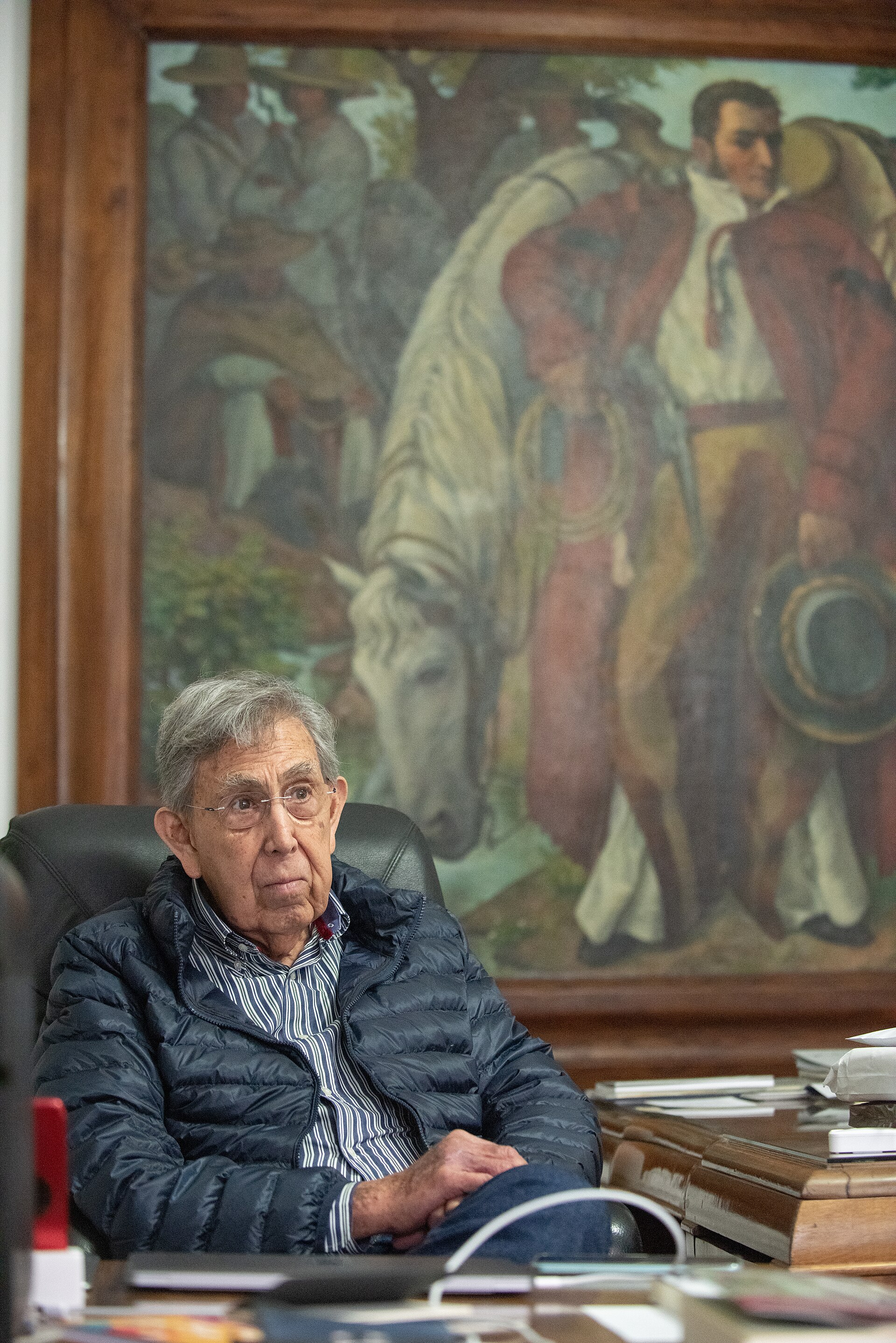 Cuauhtémoc Cárdenas, with gray hair and glasses, sitting in his office in front of a large painting of horsemen