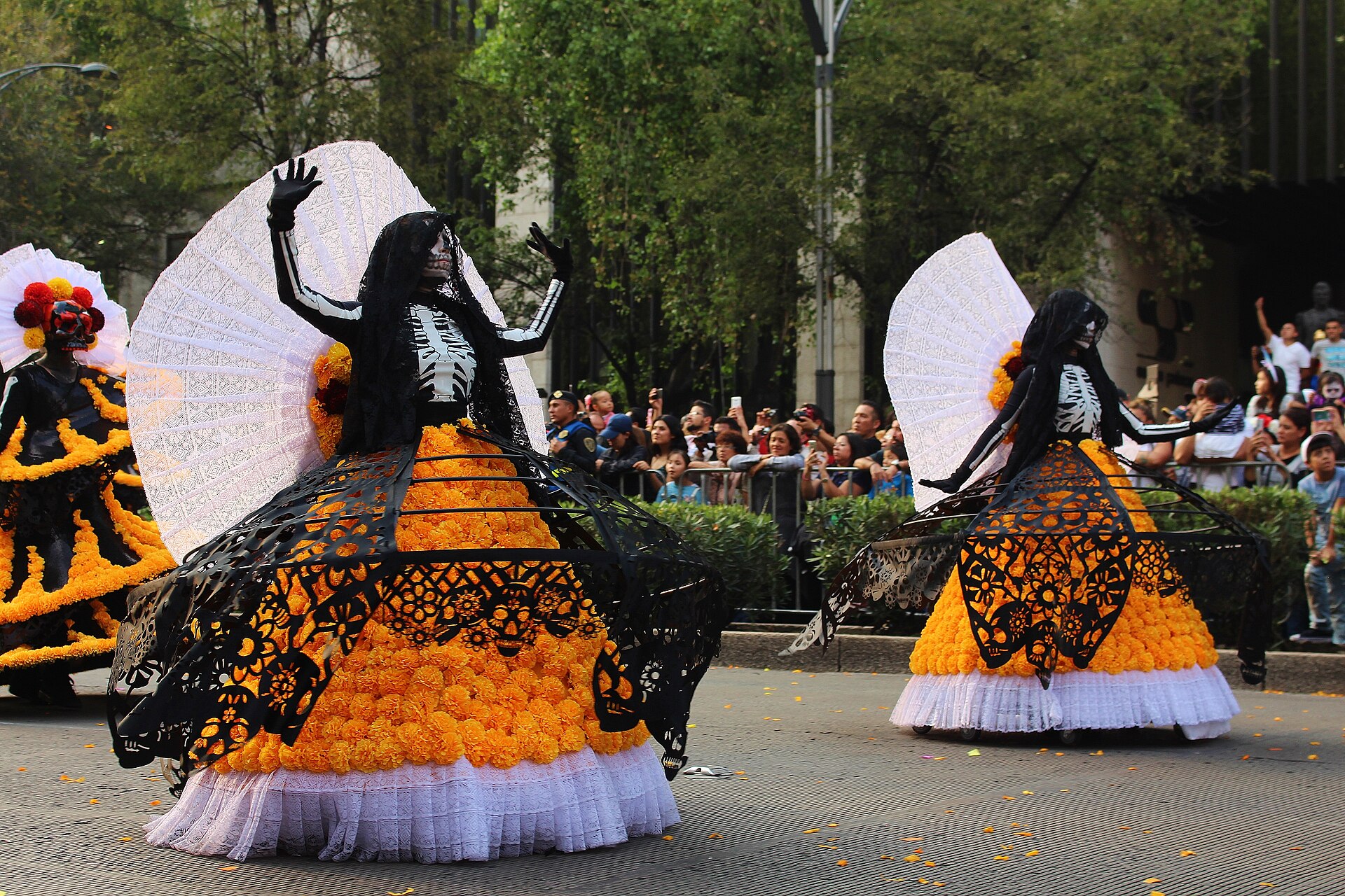 Two catrinas with huge skirts of cempasúchil marigolds and white fans parade along Paseo de la Reforma while spectators take photos
