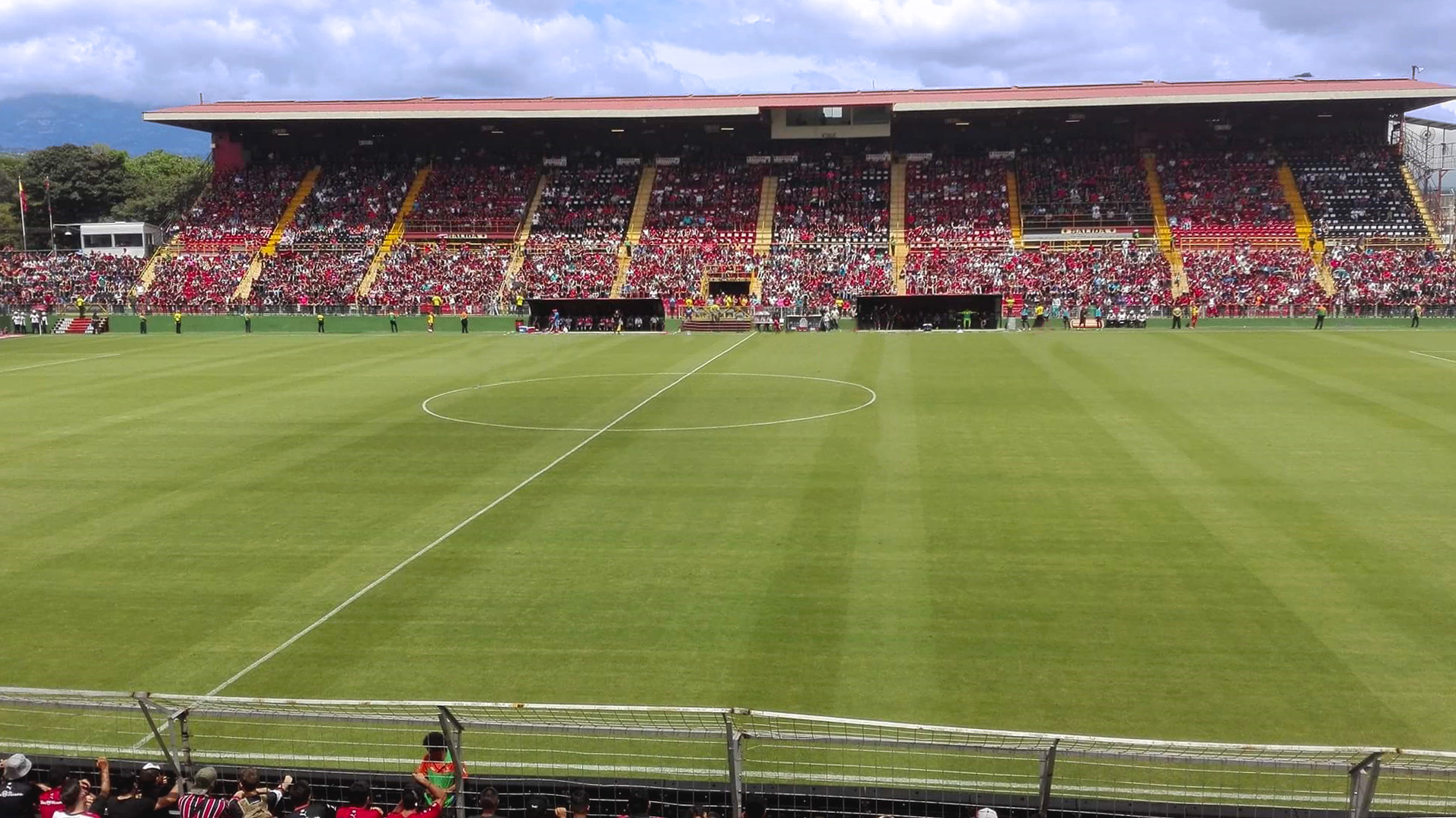 Estadio de fútbol con la grada llena de aficionados vestidos de rojo y negro y el césped verde en primer plano