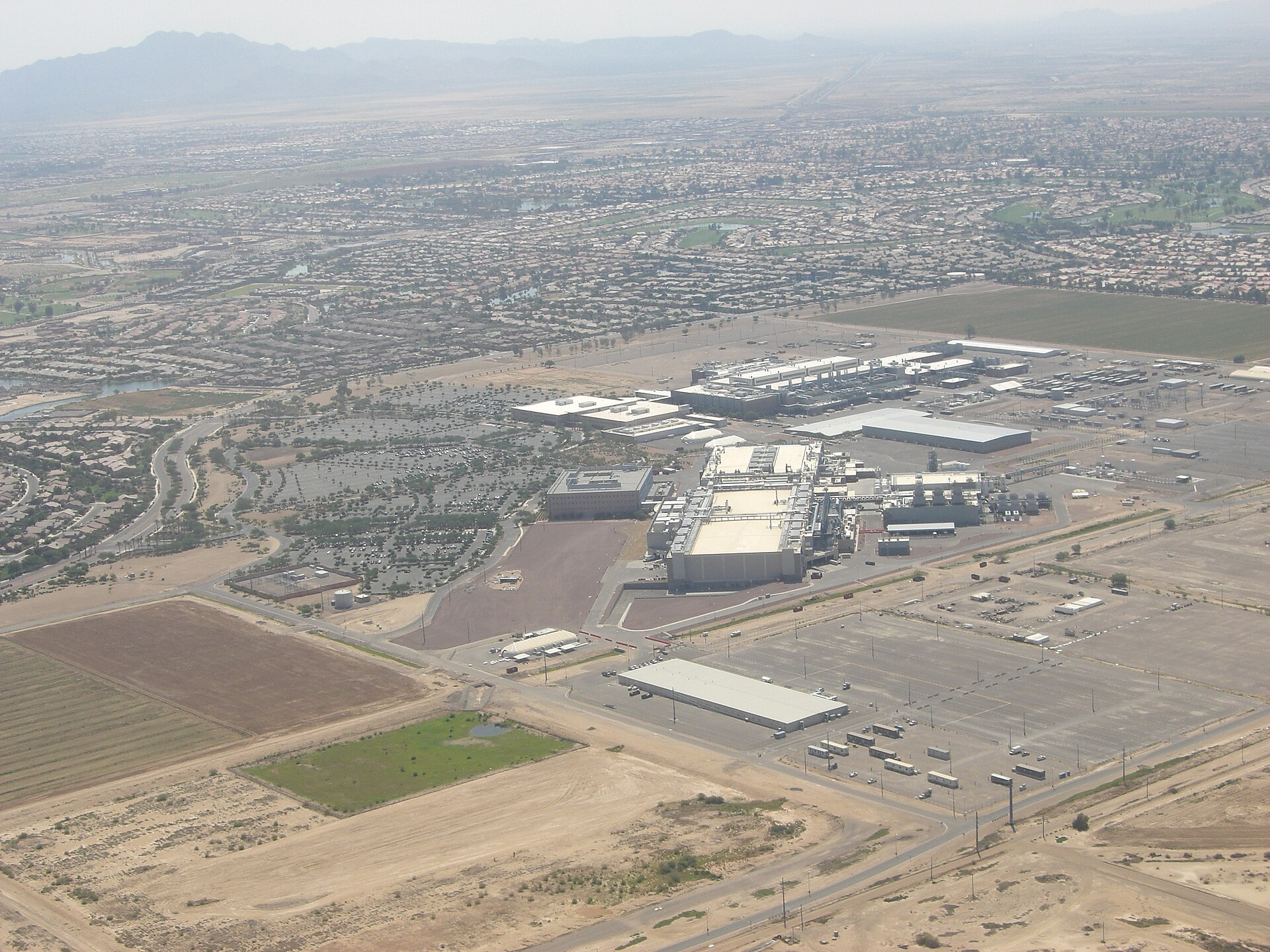 Aerial view of a large complex of white factories in the middle of the desert, with neighborhoods behind it and mountains in the background