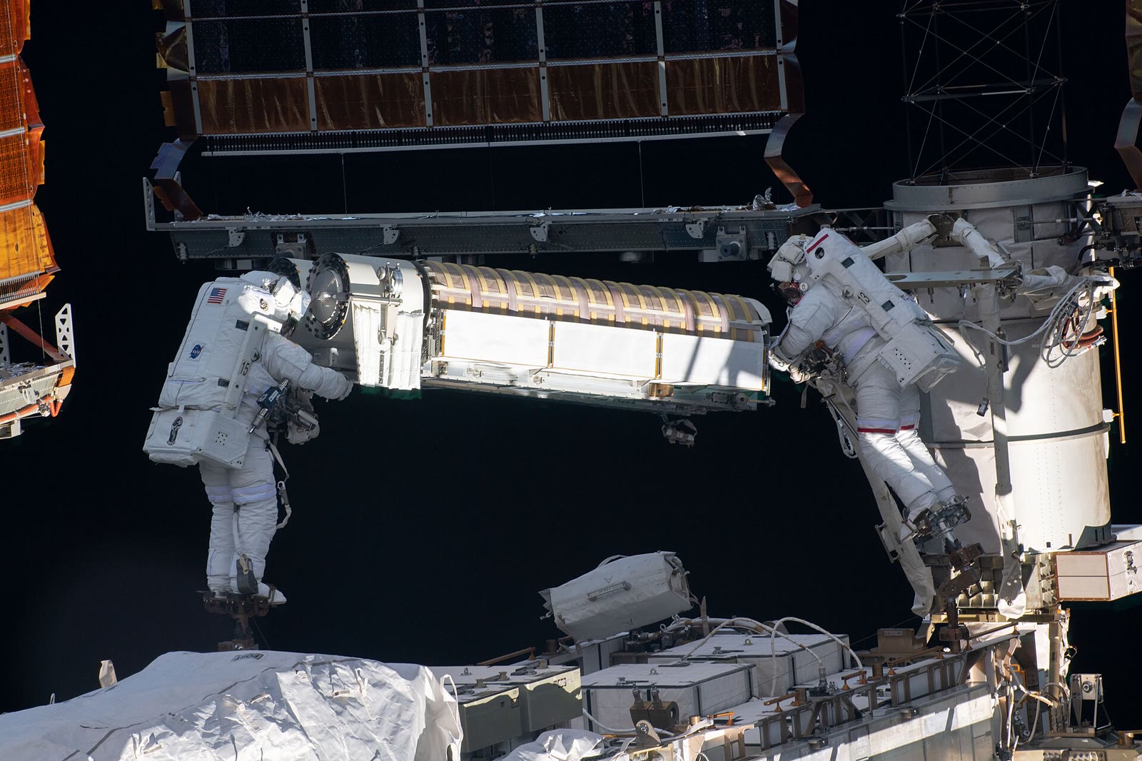Two astronauts in white spacesuits installing a rolled-up solar panel on the International Space Station