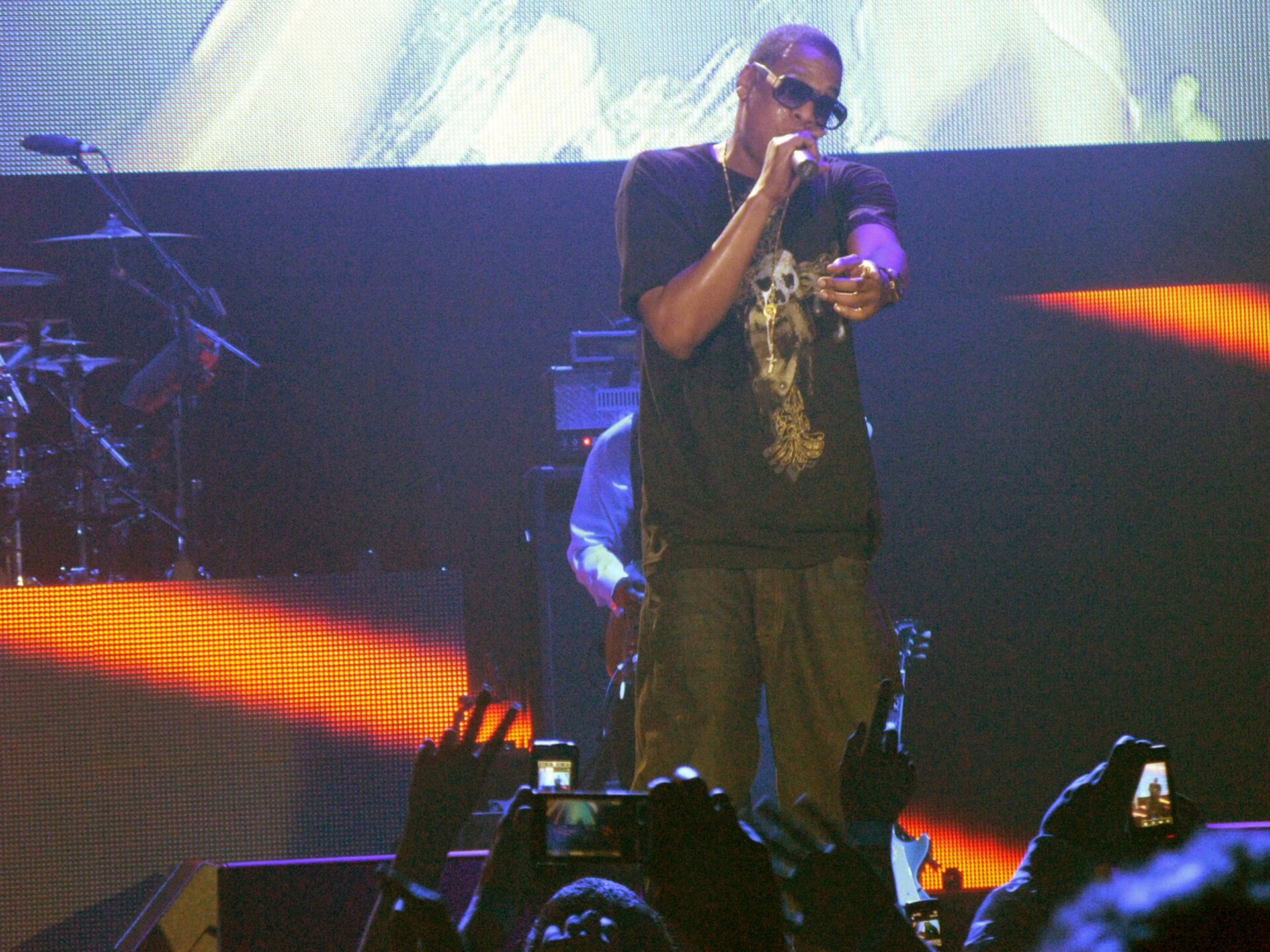 Jay-Z in dark sunglasses and a black T-shirt sings into a microphone on stage while the crowd films him with their phones