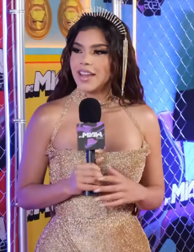 Kenia Os con un vestido dorado y una corona, hablando a un micrófono en una alfombra de premios