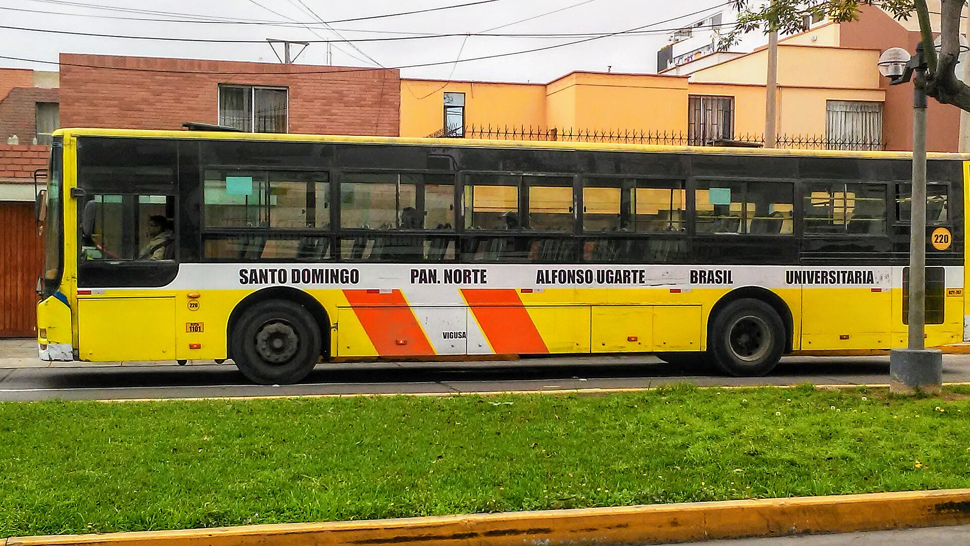A yellow city bus with orange stripes driving along an avenue in Lima, with houses behind it