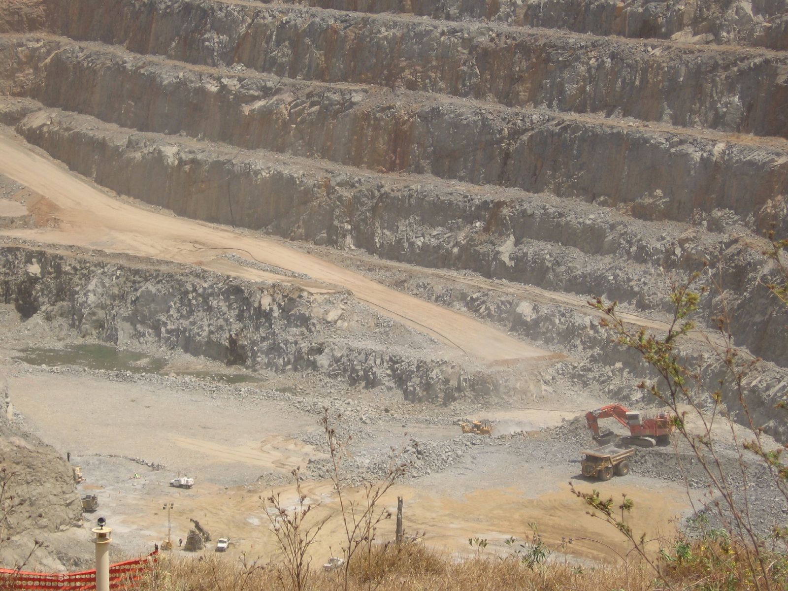 An open-pit mine with stepped walls of gray rock, and in the background a red excavator loading a giant truck