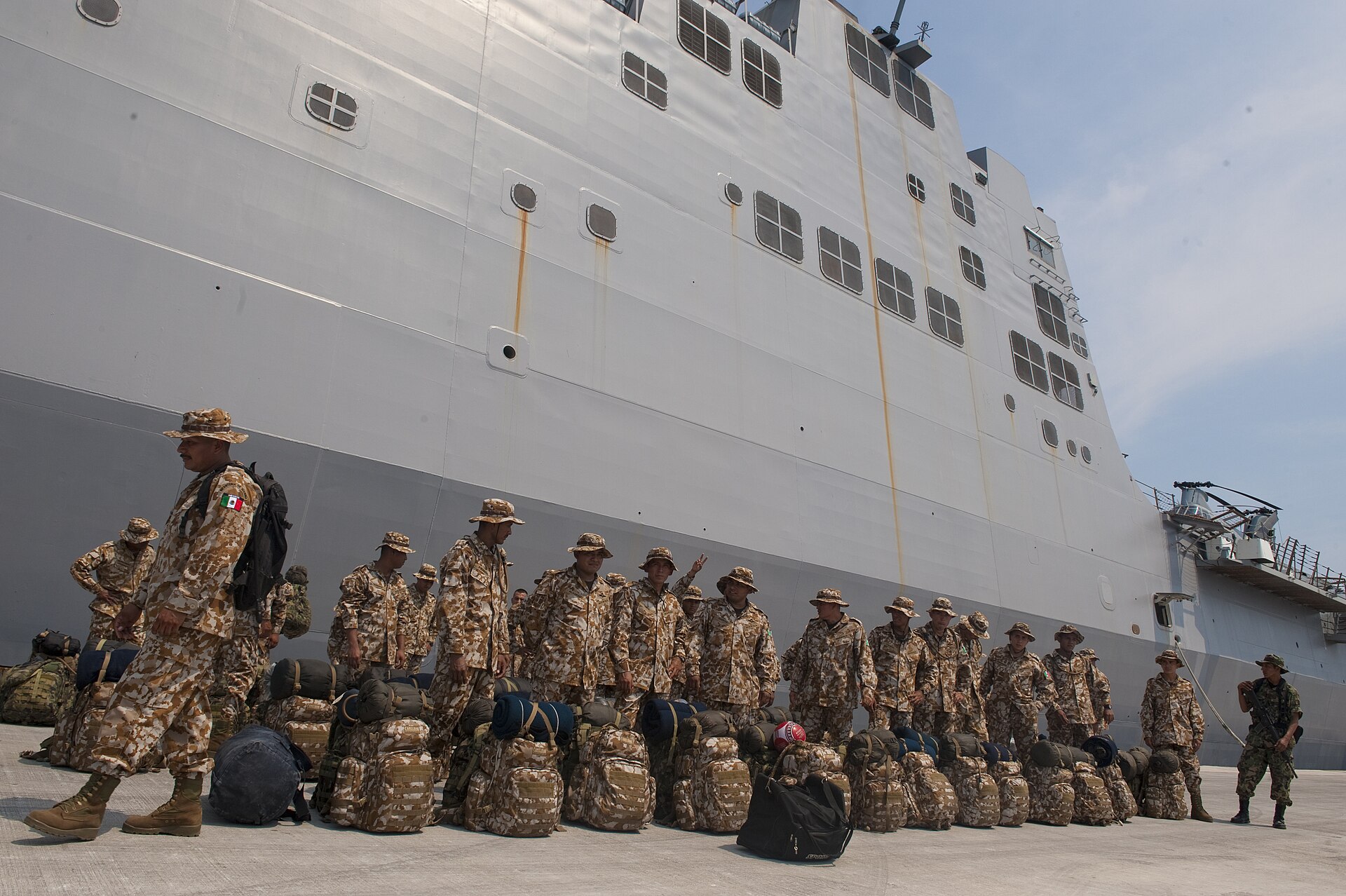 A group of Mexican marines in camouflage uniforms with backpacks next to the grey hull of a large ship