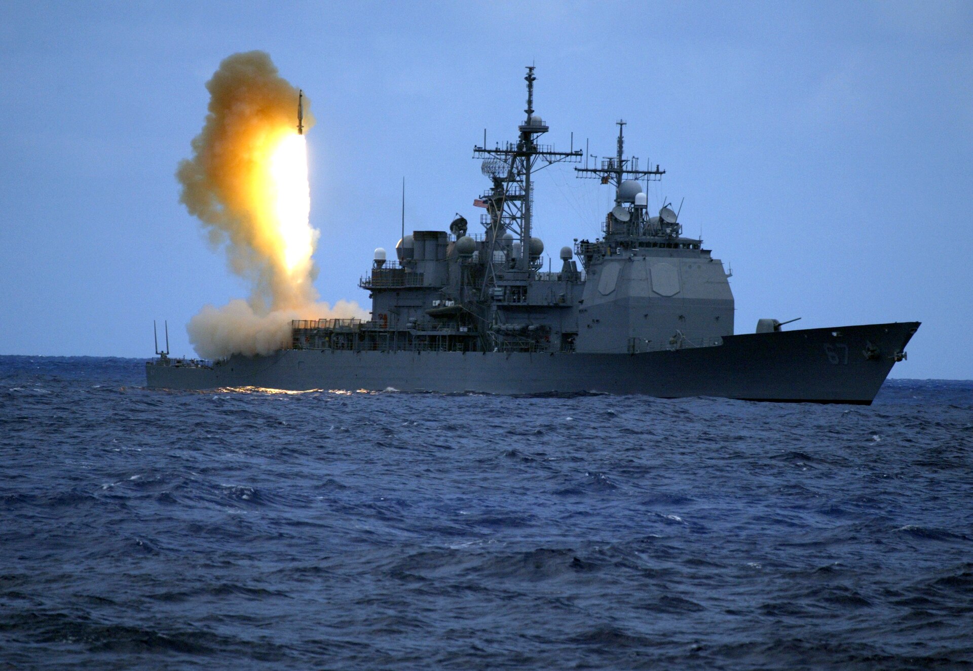 A gray warship on the open sea launches a missile that leaves a column of fire and smoke