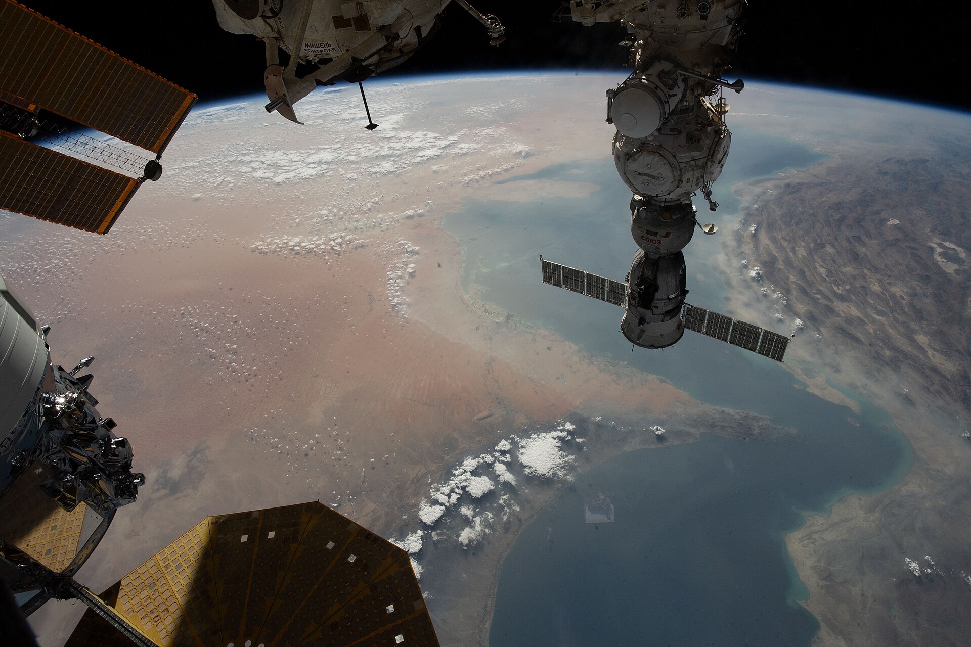 The Strait of Hormuz seen from space, with blue sea between two desert coasts and parts of the International Space Station in the foreground