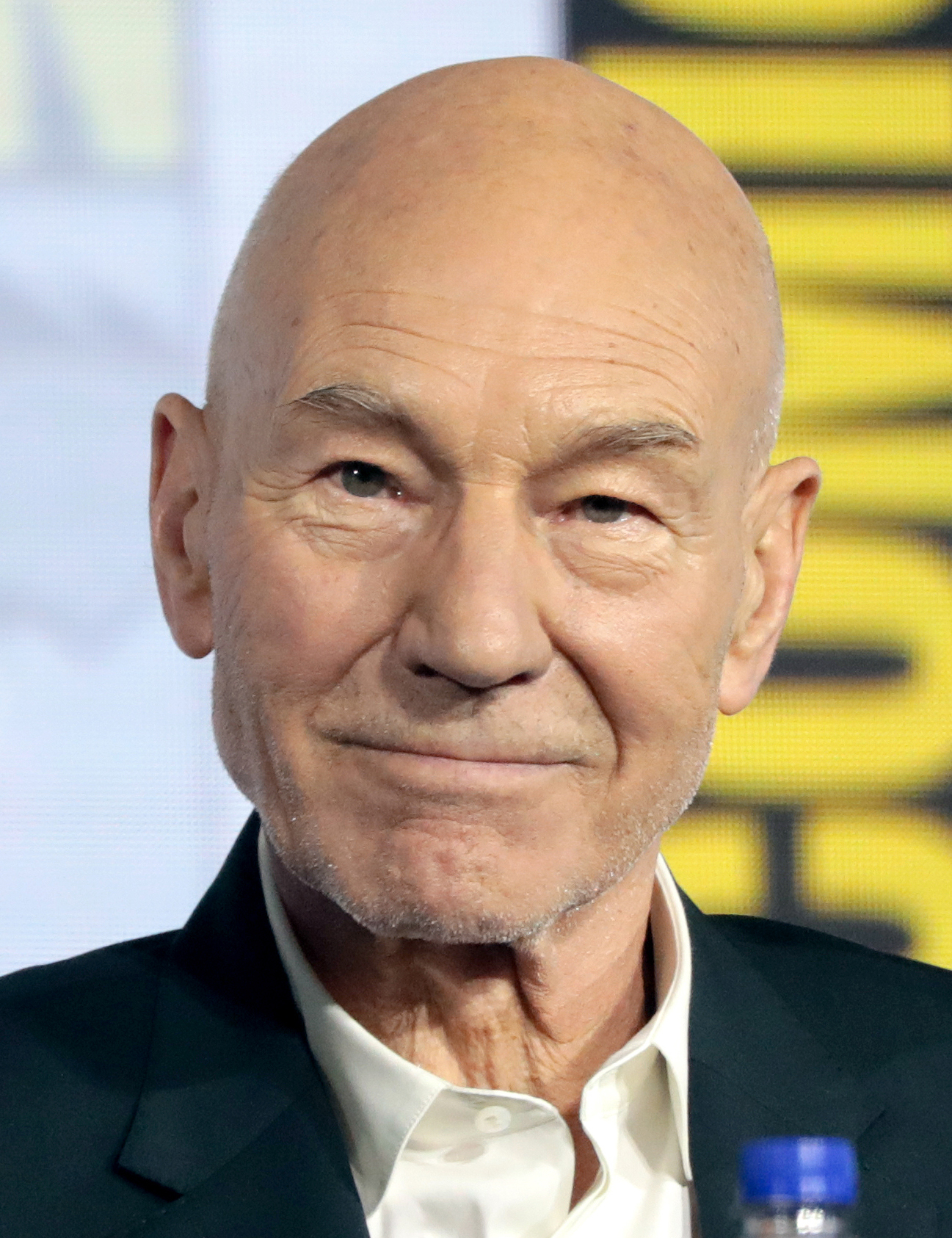 Patrick Stewart sorrindo, de terno escuro e camisa branca, em um painel com fundo amarelo