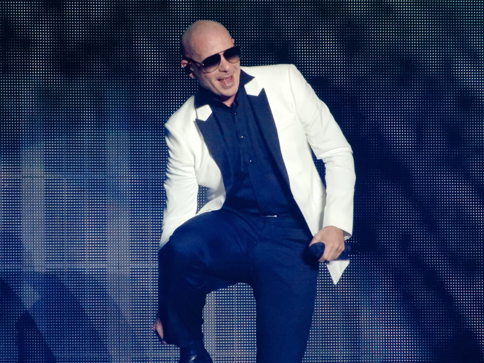 Pitbull de óculos escuros, paletó branco e calça azul-escura canta com o microfone na mão em um palco com luzes azuis