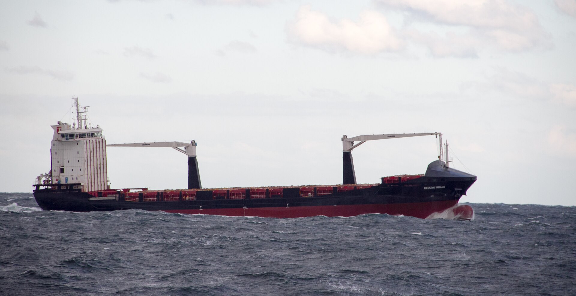 Un barco portacontenedores negro y rojo con dos grúas navegando por un mar agitado bajo un cielo nublado