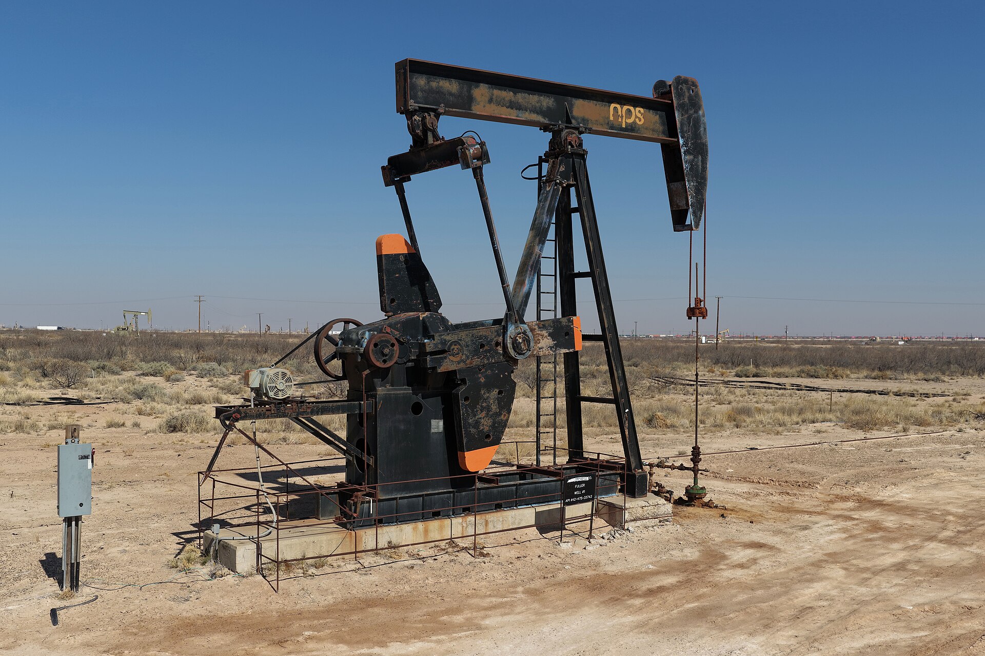 Bomba de extracción de petróleo, negra con detalles naranjas, en un campo seco bajo un cielo azul