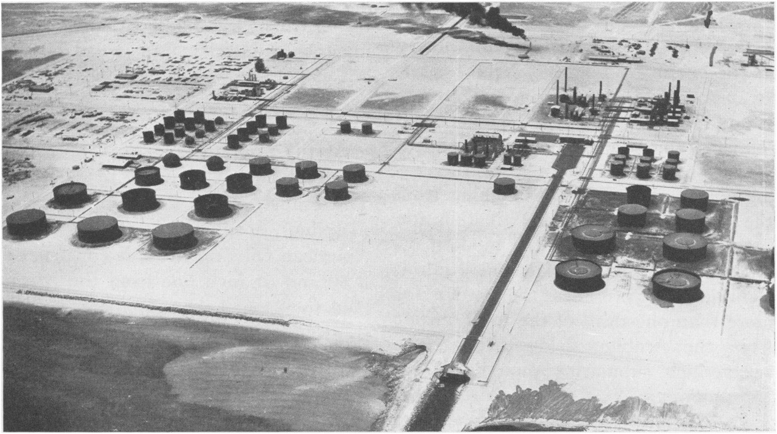 Foto antigua en blanco y negro de la refinería de Ras Tanura, con tanques y chimeneas junto al mar