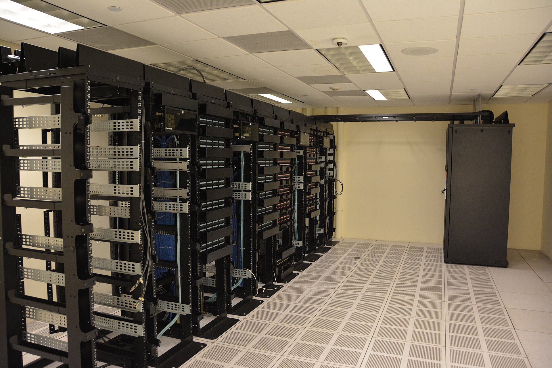 Rows of black cabinets full of cables and servers in a room with a raised floor and a white ceiling