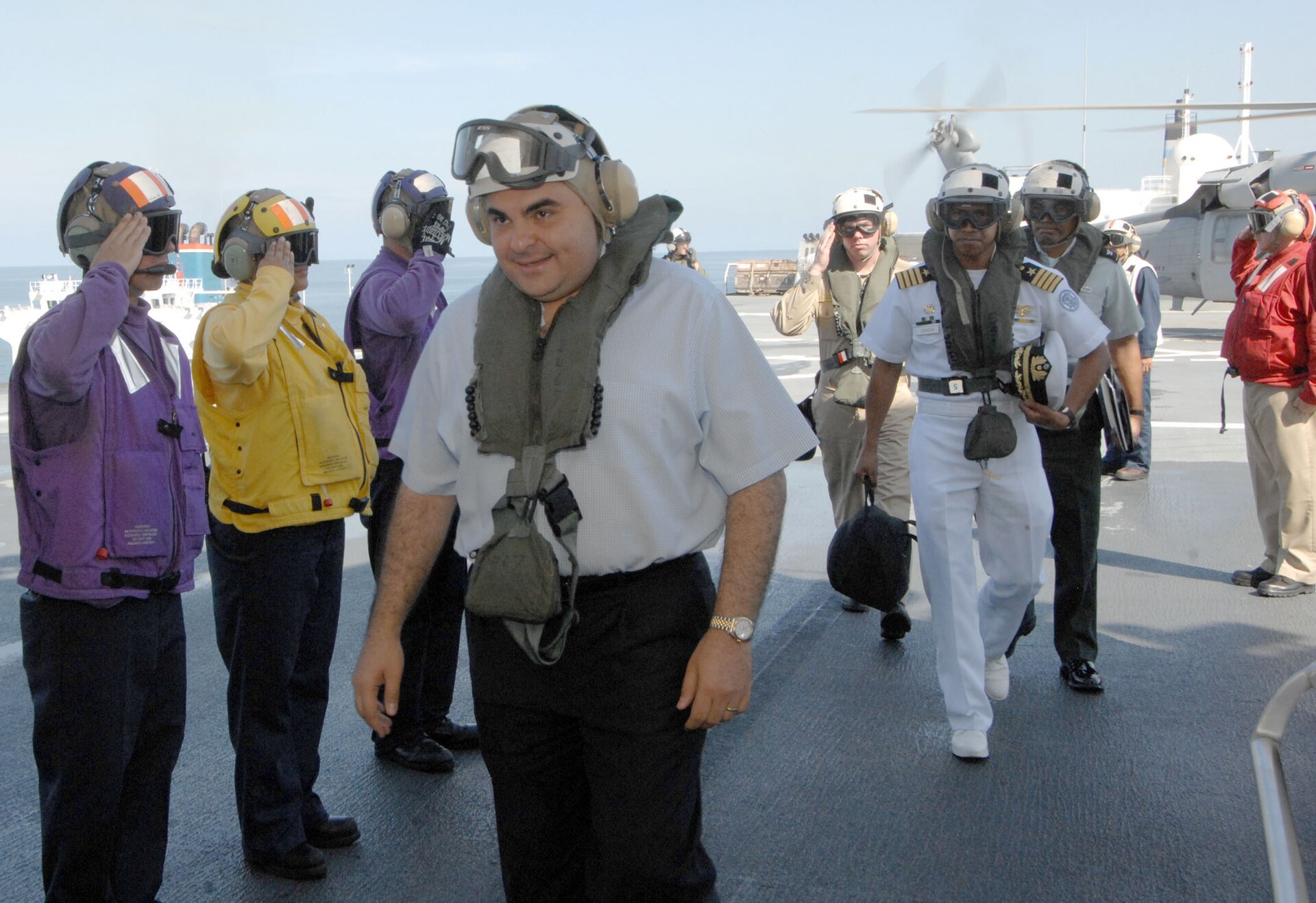 Elías Antonio Saca, con camisa clara y chaleco salvavidas, camina sonriendo por la cubierta de un barco mientras marinos lo saludan