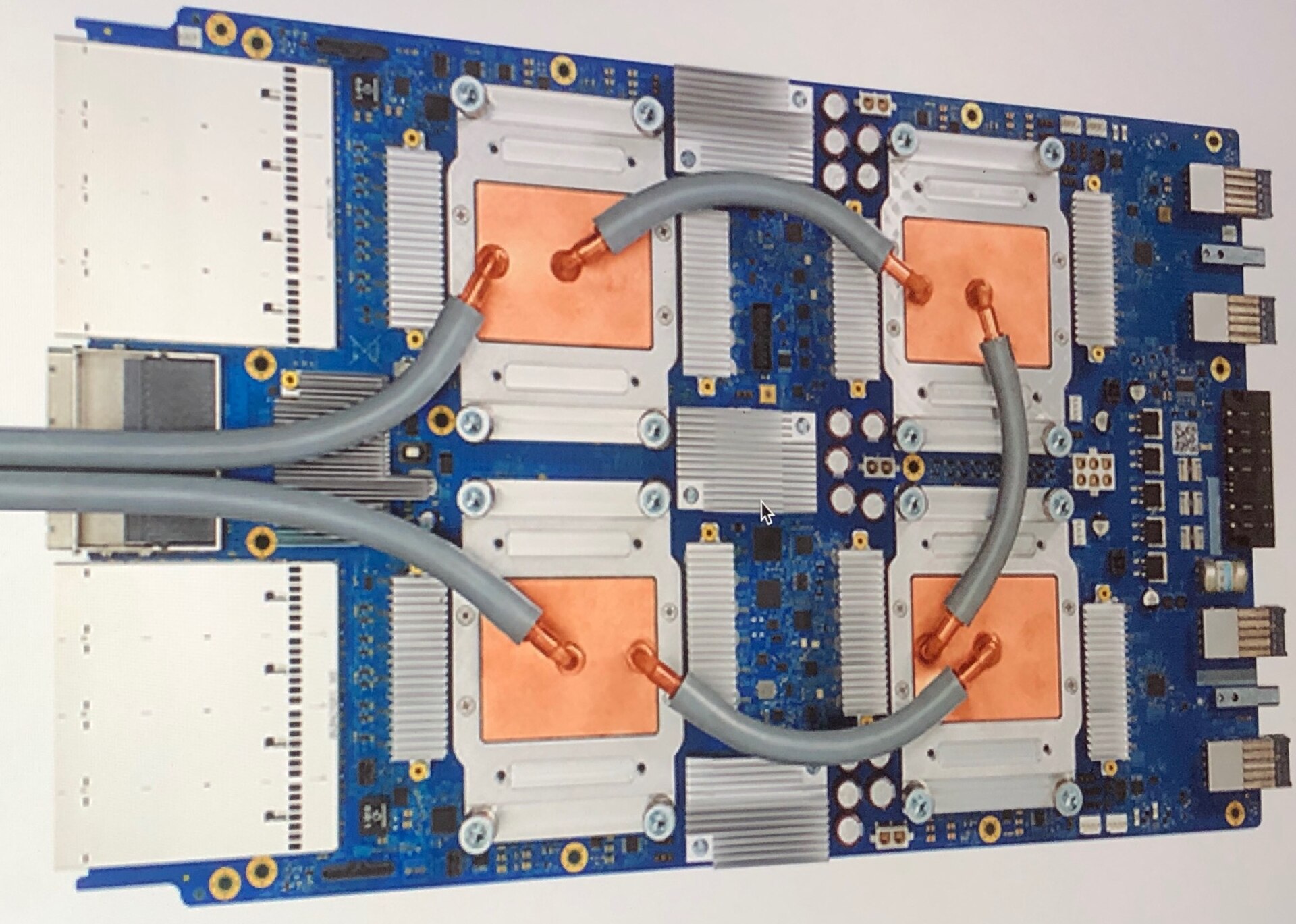 A blue circuit board with four chips covered by copper blocks and connected by gray cooling tubes