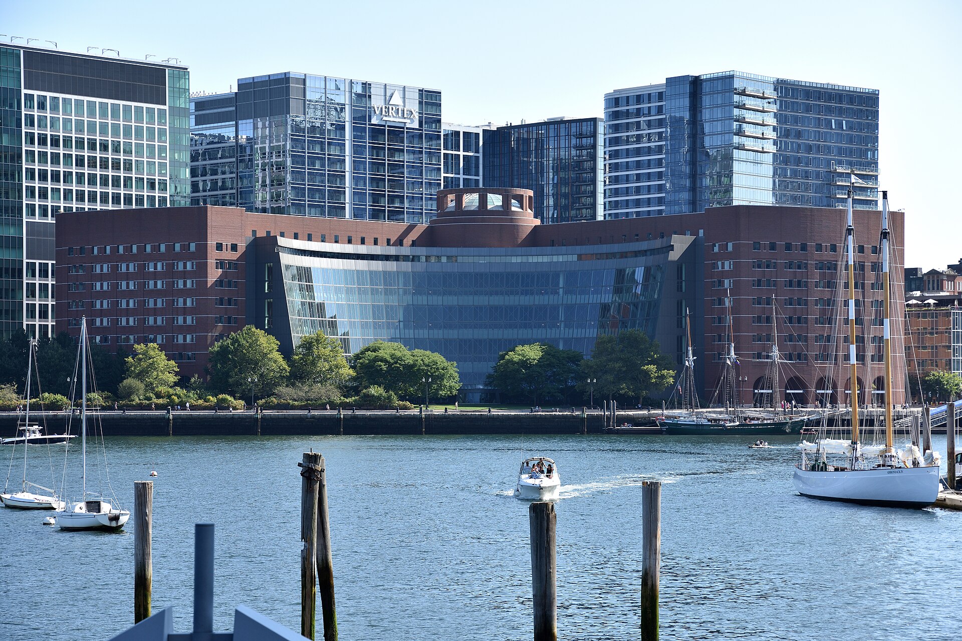A large red-brick building with a curved glass facade next to Boston Harbor, with sailboats and a motorboat on the water